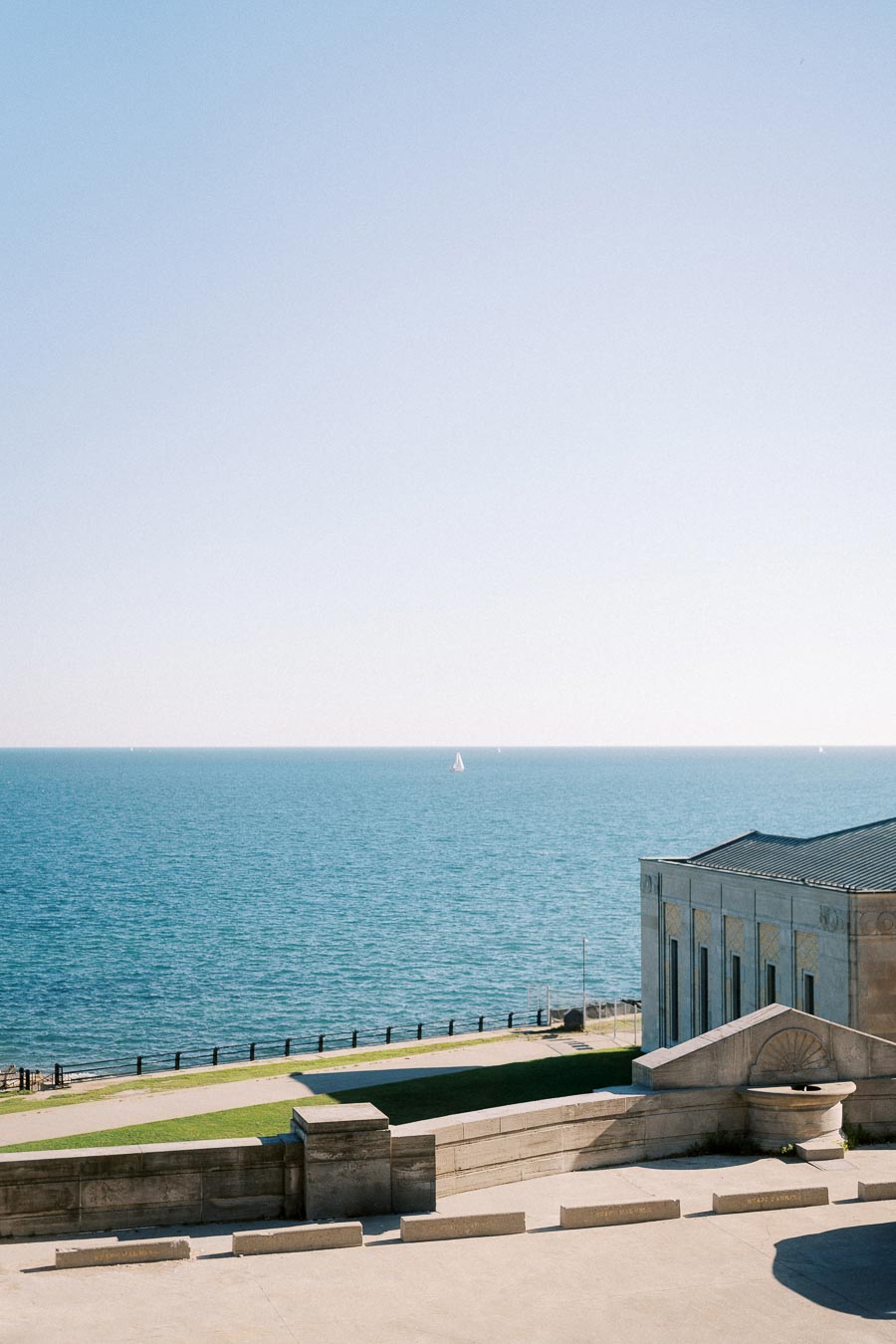 Serene coastal view with a historic building overlooking the ocean and a sailboat in the distance under a clear blue sky.