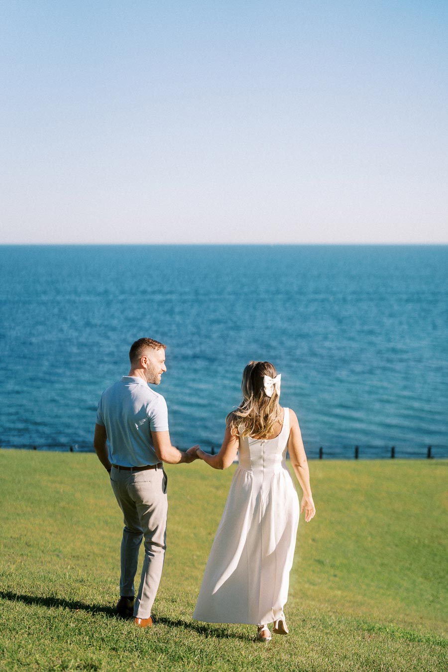 A couple holding hands and walking on green grass toward the ocean under a clear blue sky.