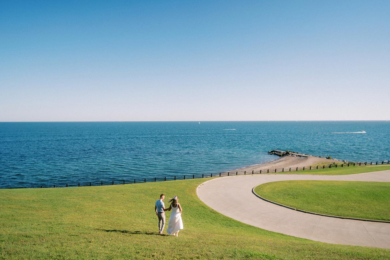A couple walks hand in hand on a grassy hill overlooking a serene ocean view under a clear blue sky.