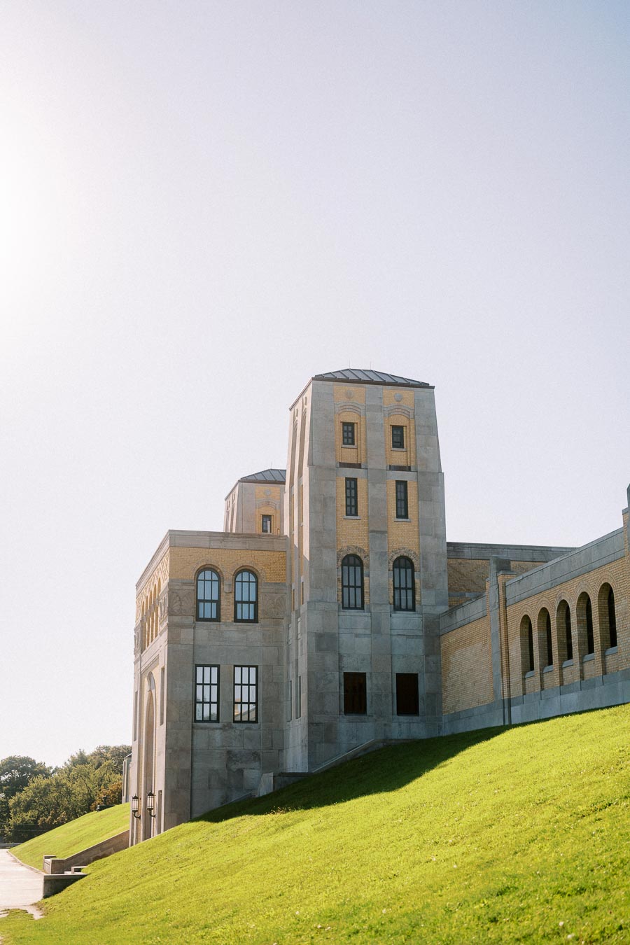 Historic stone building with arched windows against a clear sky, surrounded by a green grassy hill, under bright sunlight.
