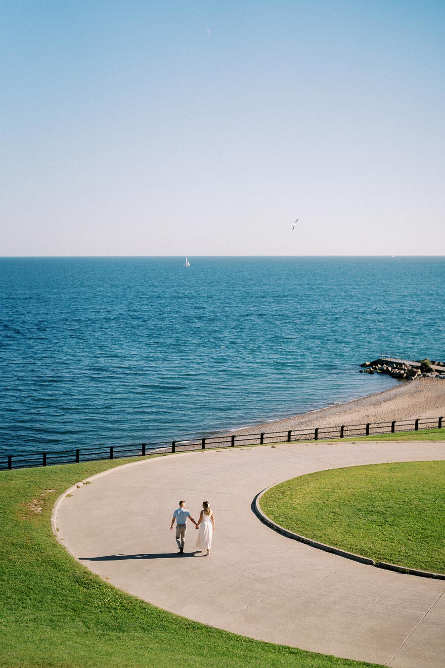 Couple holding hands walking along a curved pathway beside a tranquil ocean on a clear day, with a sailboat in the distance and seagulls flying overhead, capturing a serene beachside moment.