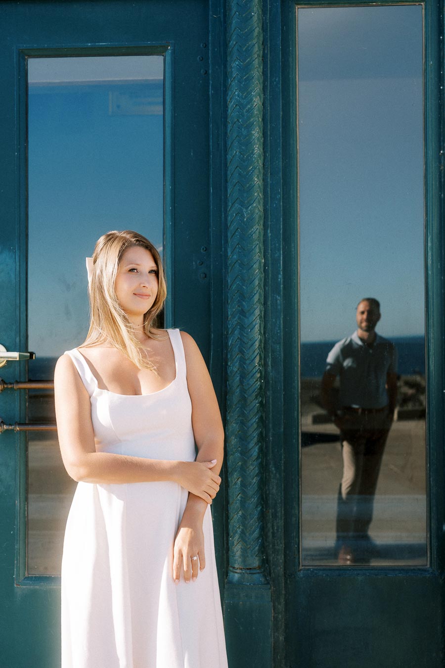 A woman in a white dress stands in front of blue doors, with a man visible in the reflection, under a clear sky.