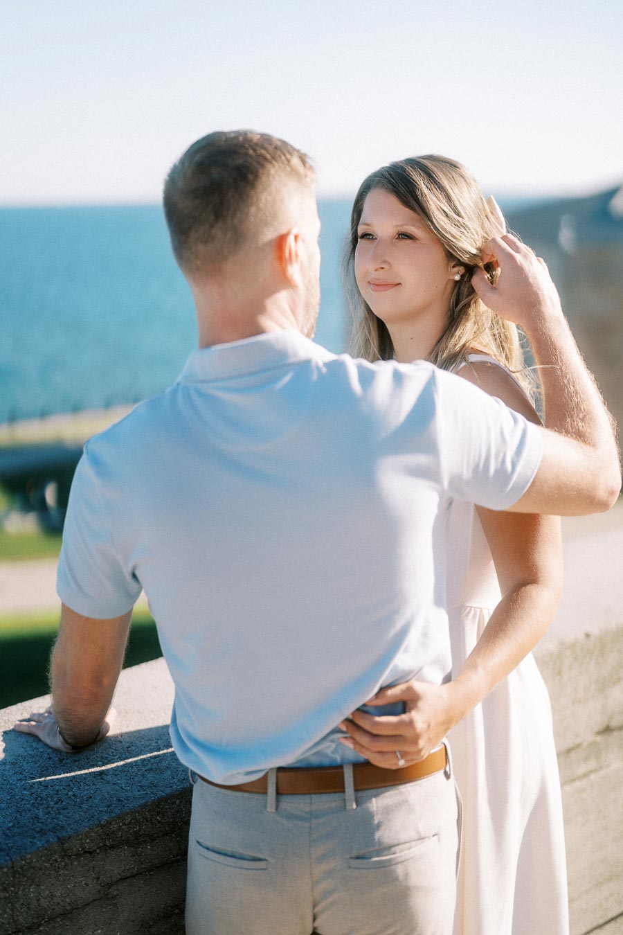 A couple sharing a tender moment by the seaside, with the woman smiling softly as they stand close together, bathed in sunlight, overlooking the ocean.