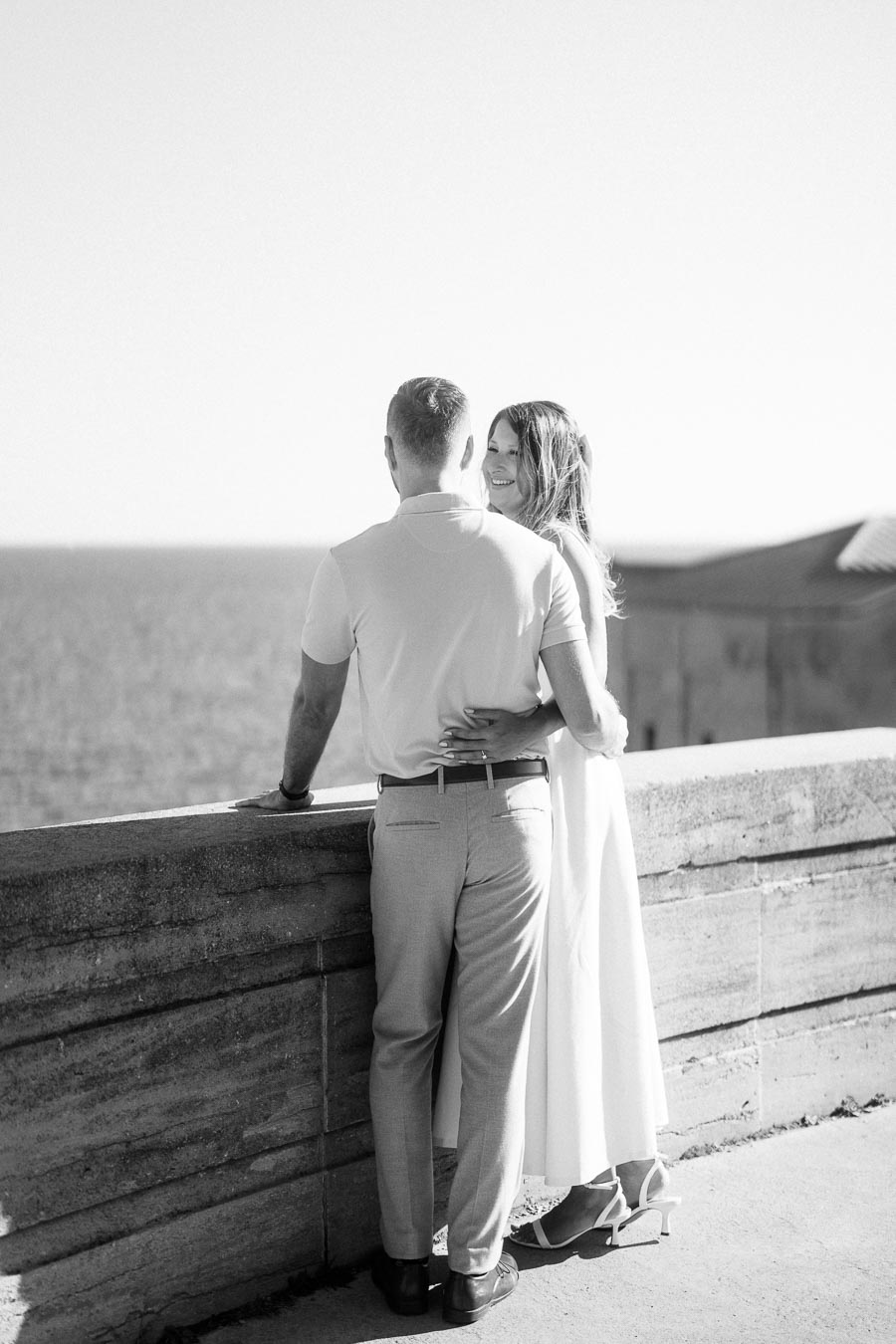 A couple embraces while gazing at the ocean from a stone balcony, captured in black and white.