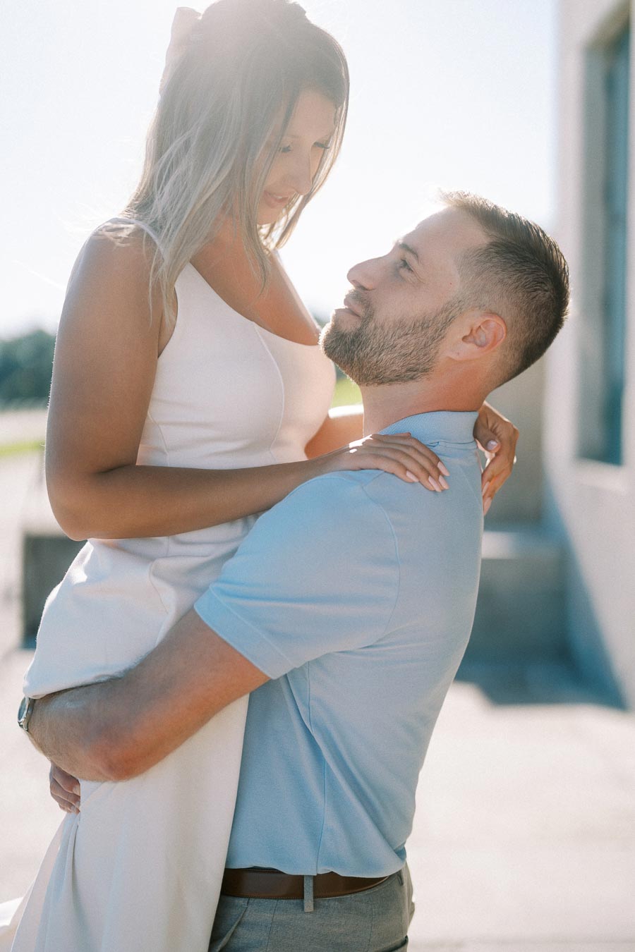 A couple in love sharing an intimate moment outdoors, with the man lifting the woman while they gaze into each other's eyes, dressed casually under soft natural lighting.