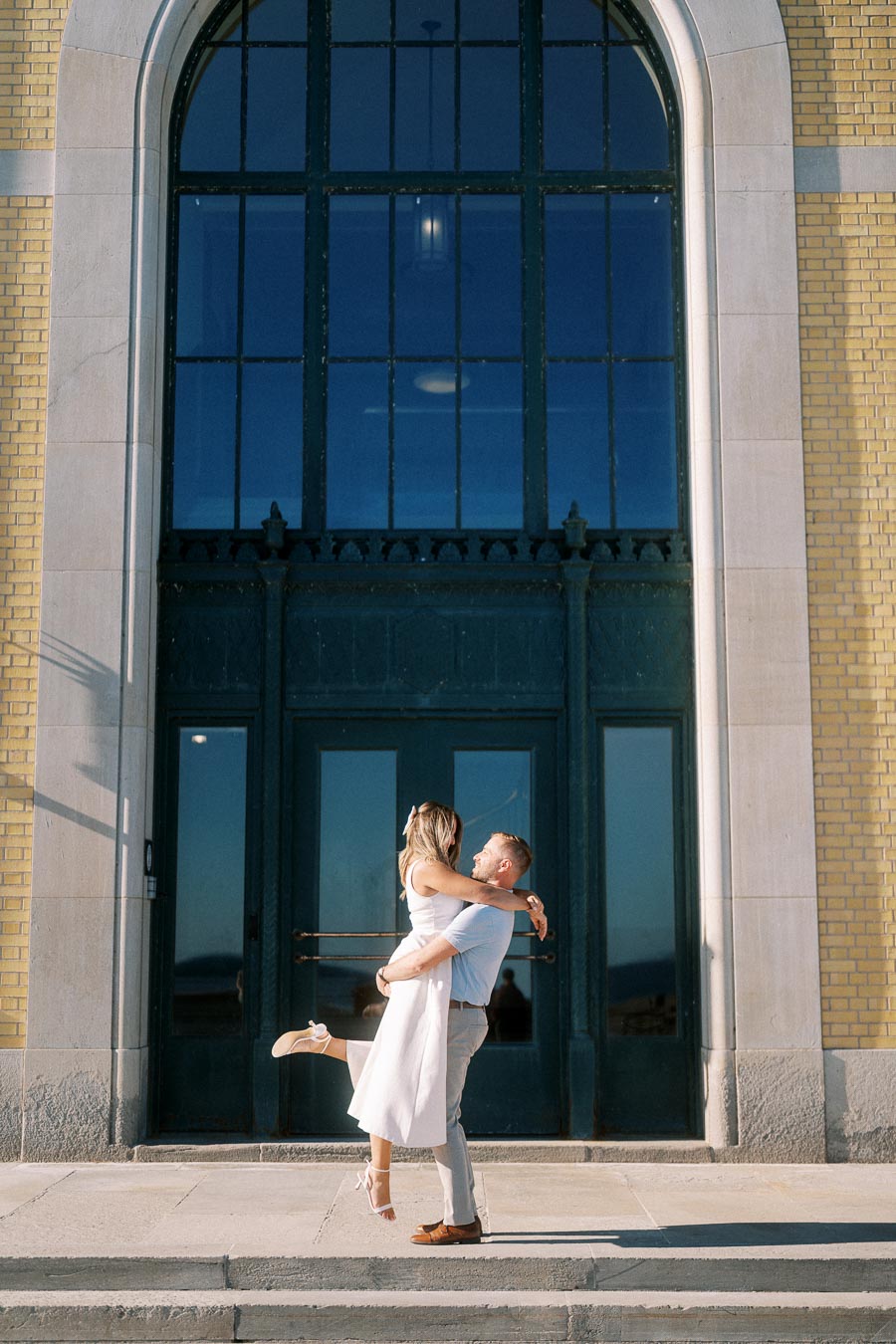 A couple embracing joyfully in front of a grand building with large arched windows and yellow brick facade.