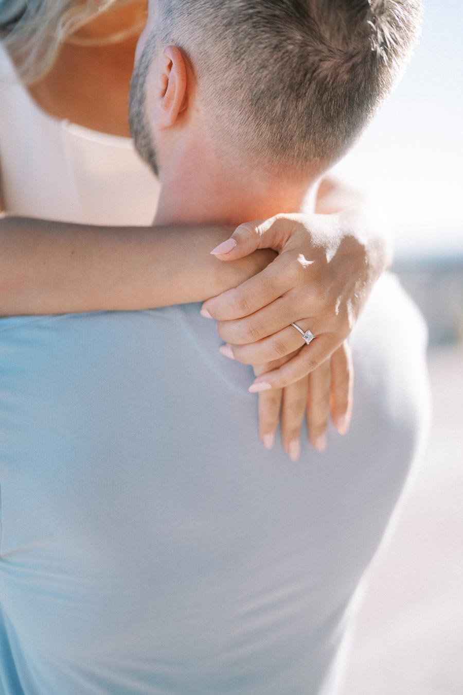 A woman with an engagement ring embraces a man in a light blue shirt, highlighting a romantic moment.