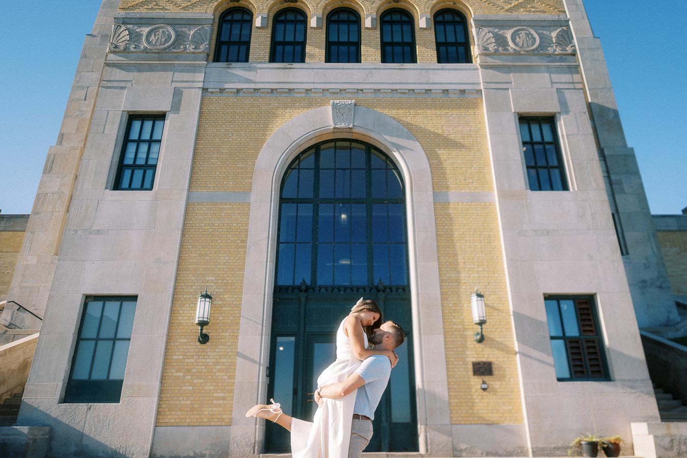A couple embracing in front of a historic building with large arched windows and ornate architectural details, set against a clear blue sky.