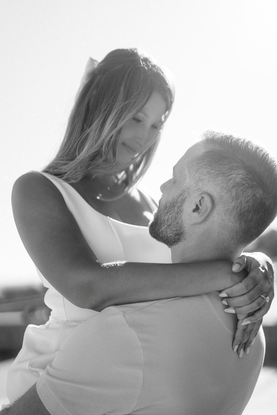 Black and white image of a couple embracing outdoors, showcasing a woman in a white dress smiling as she wraps her arms around a man.