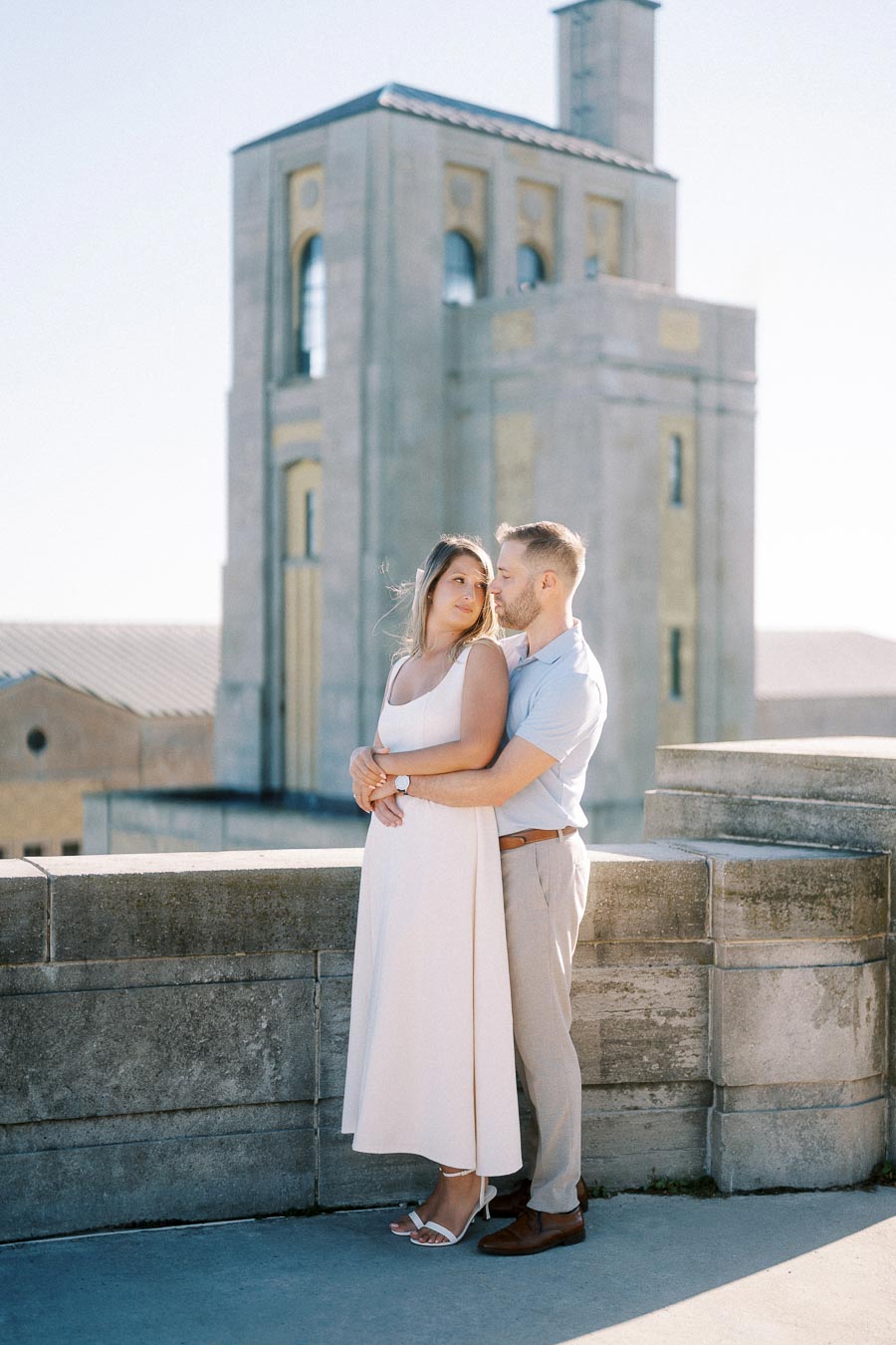 A couple embracing and smiling in front of a historic stone building under clear blue skies.