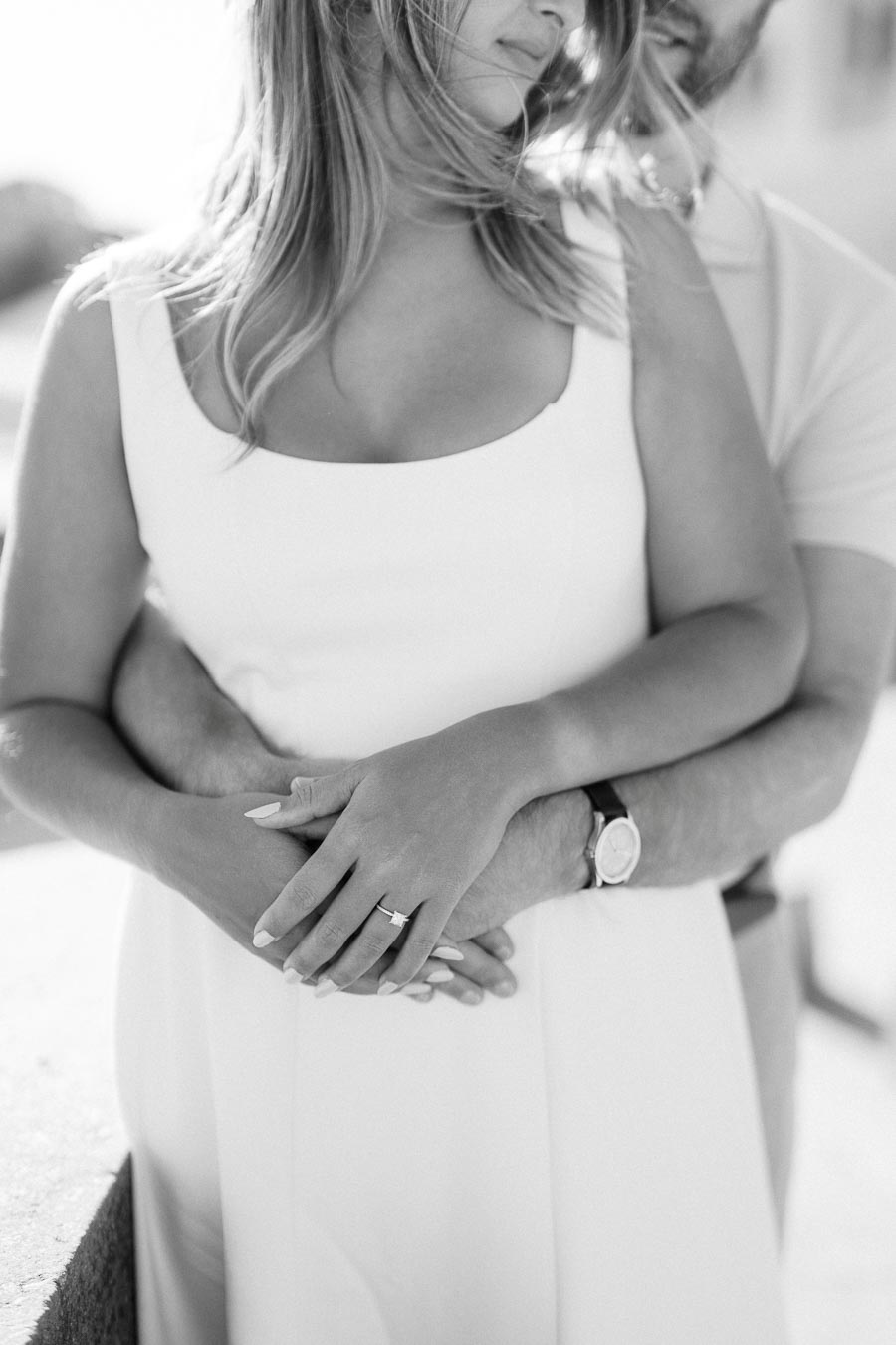 Black and white photo of a couple embracing, highlighting a woman's engagement ring on her hand against a white dress, with a man's arms wrapped around her waist, capturing a romantic moment.