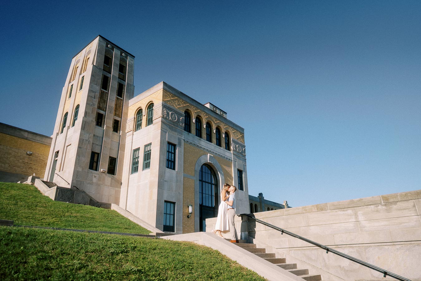 Couple embracing on steps in front of historic stone building under clear blue sky, surrounded by green grass.