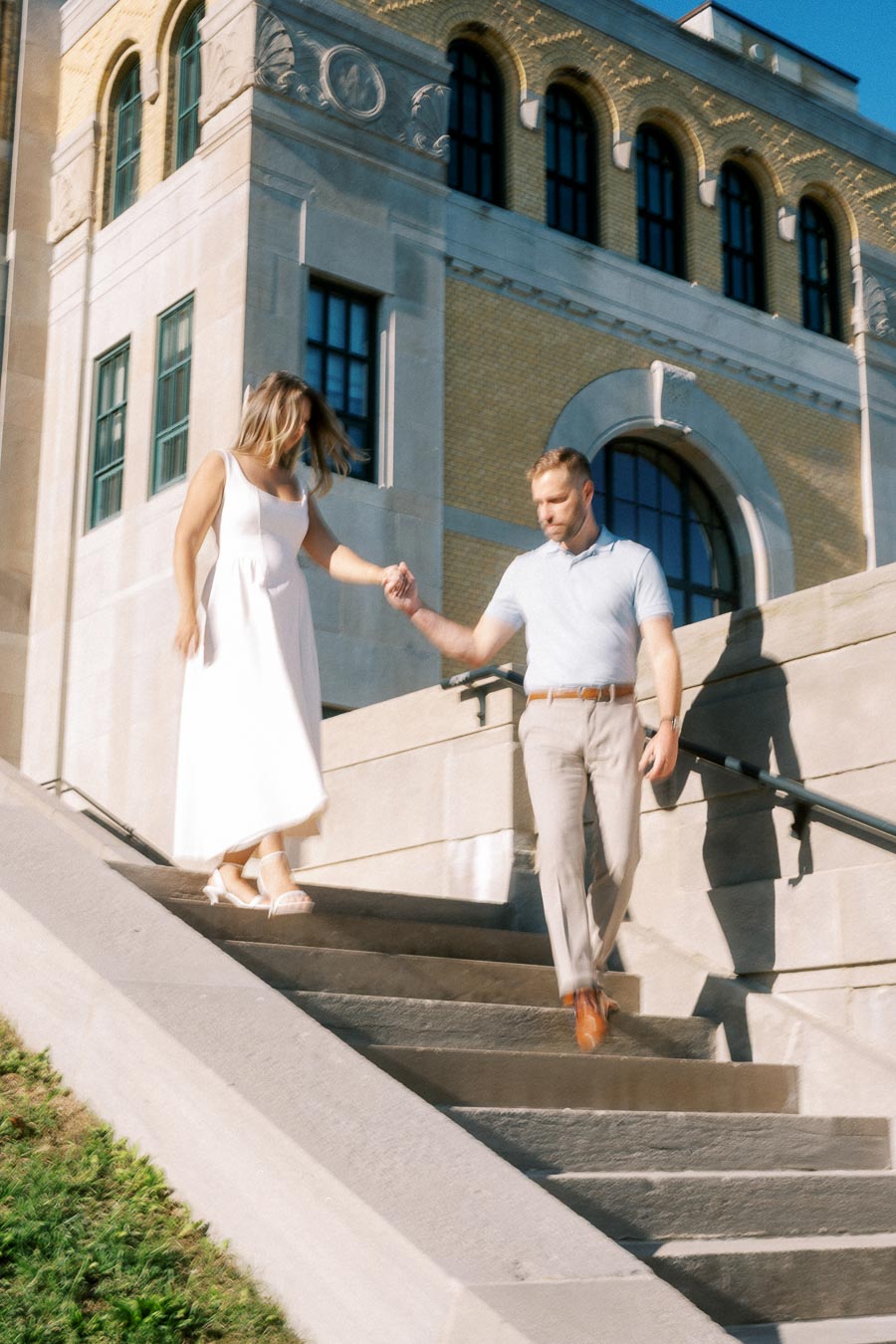A couple elegantly descends stone steps outside a historic building on a sunny day, highlighting architecture and summer fashion.