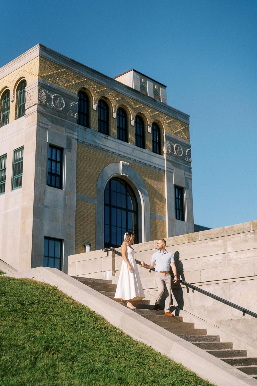 Couple walking down stone steps in front of a historic building with large arched windows and detailed architecture under a clear blue sky.