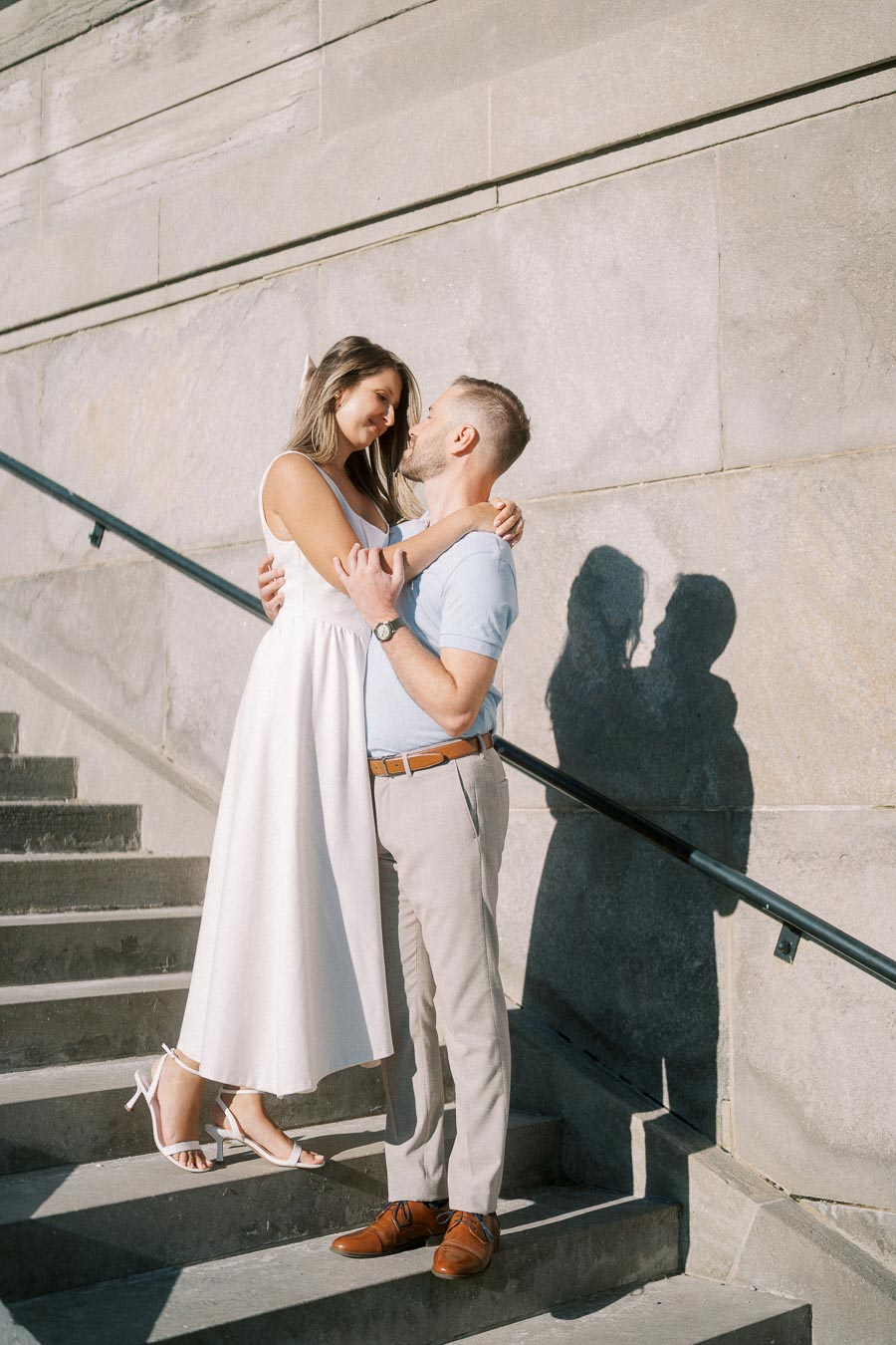 A couple embracing lovingly on stone steps, casting a shadow on a textured wall behind them, with soft sunlight enhancing the romantic atmosphere.