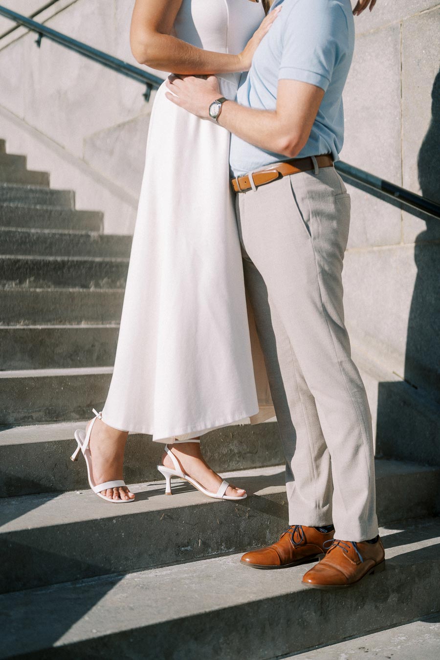 A couple standing closely together on a stone staircase, with the woman wearing a long white dress and heels, and the man in a light blue shirt and beige pants, showcasing an intimate and stylish moment.