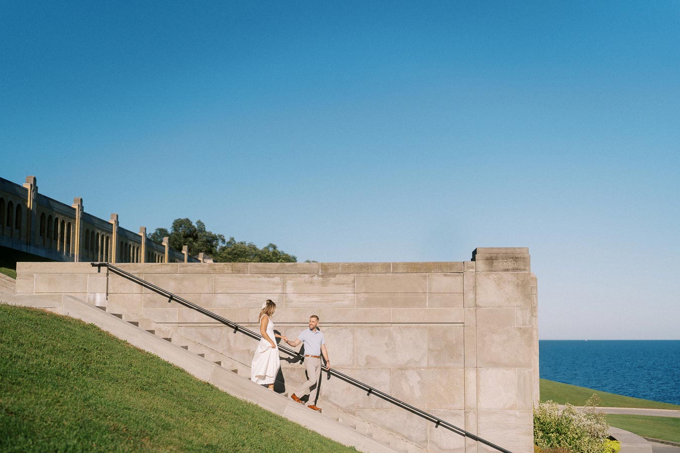 A couple walking hand in hand on concrete stairs near a park with a scenic view of the blue sea and a clear sky.