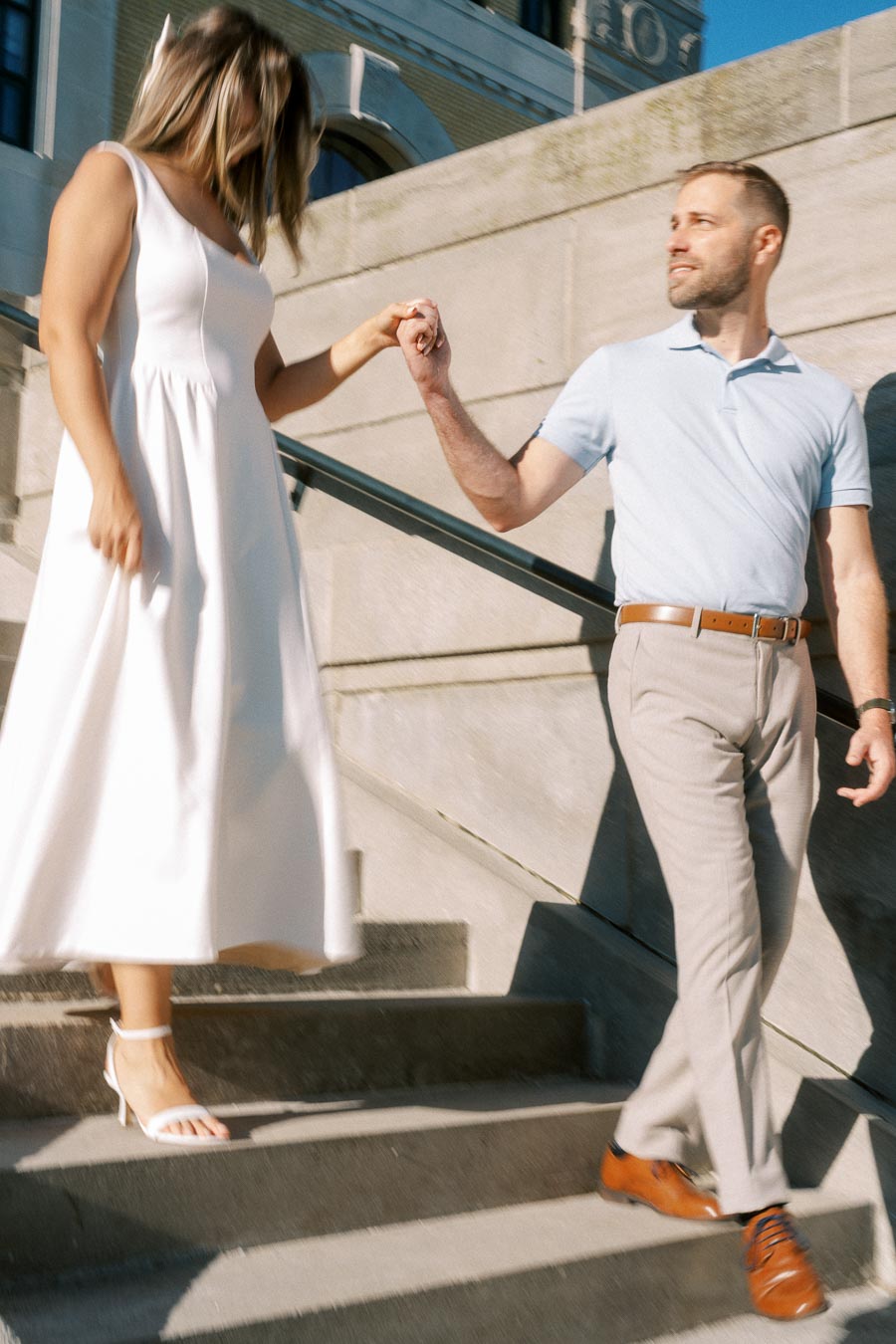 A couple walking down stone stairs, holding hands, with a woman in a white dress and a man in a light blue shirt and beige pants.