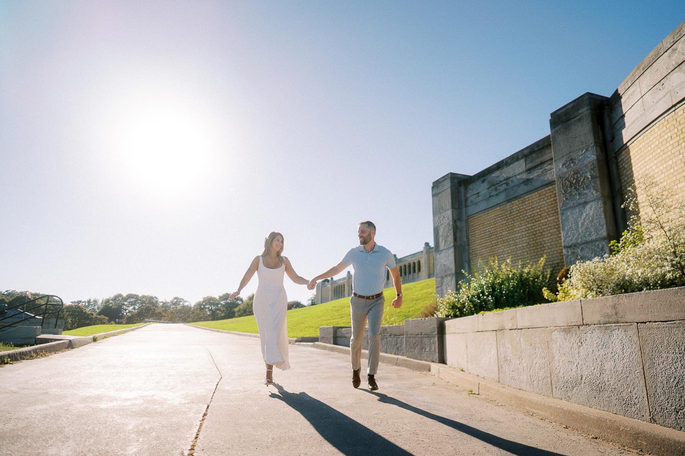 A couple holding hands while joyfully walking on a sunny day by a stone wall and greenery, with a bright blue sky in the background.