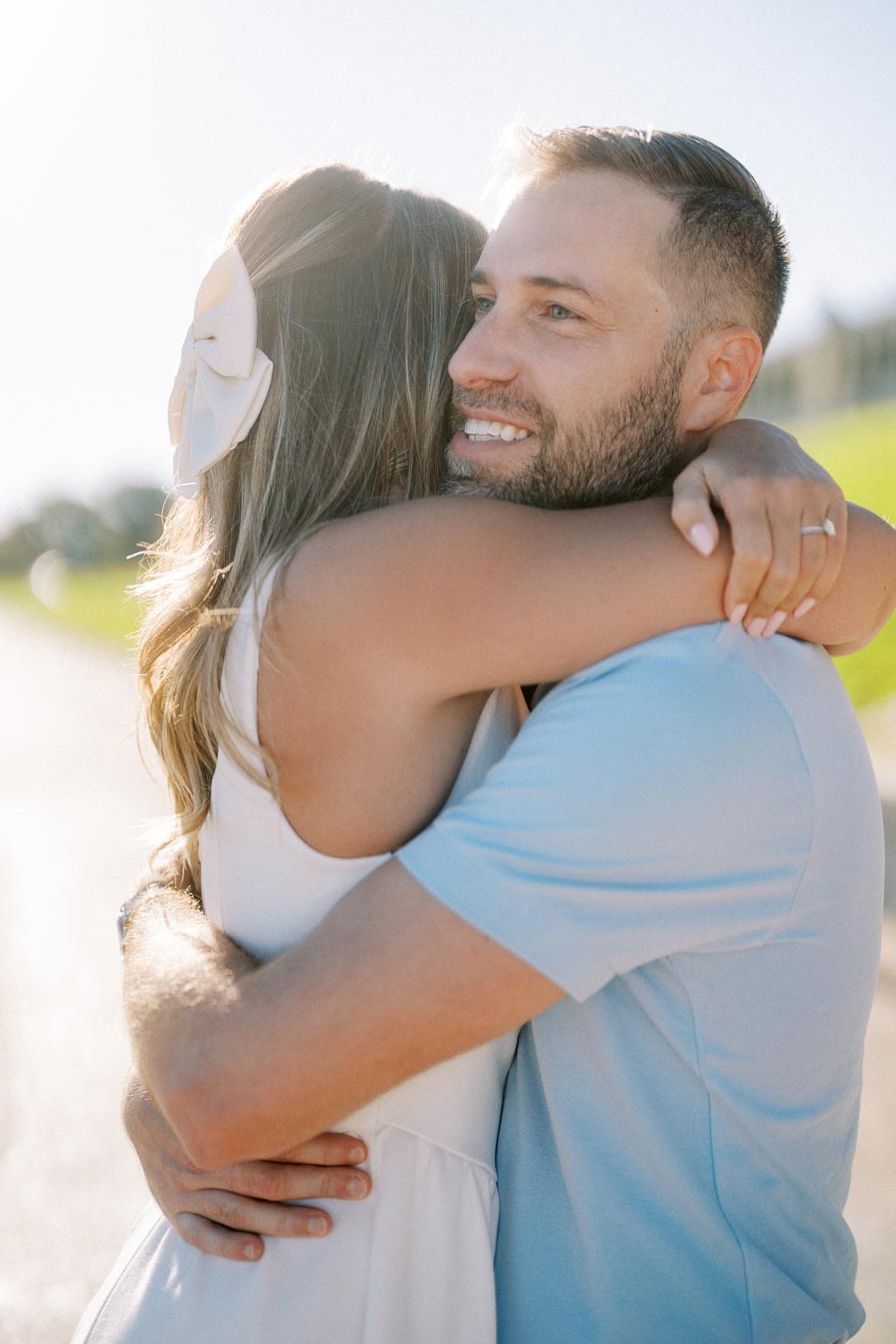 A couple embracing outdoors on a sunny day, with the man smiling and the woman with a ribbon in her hair.
