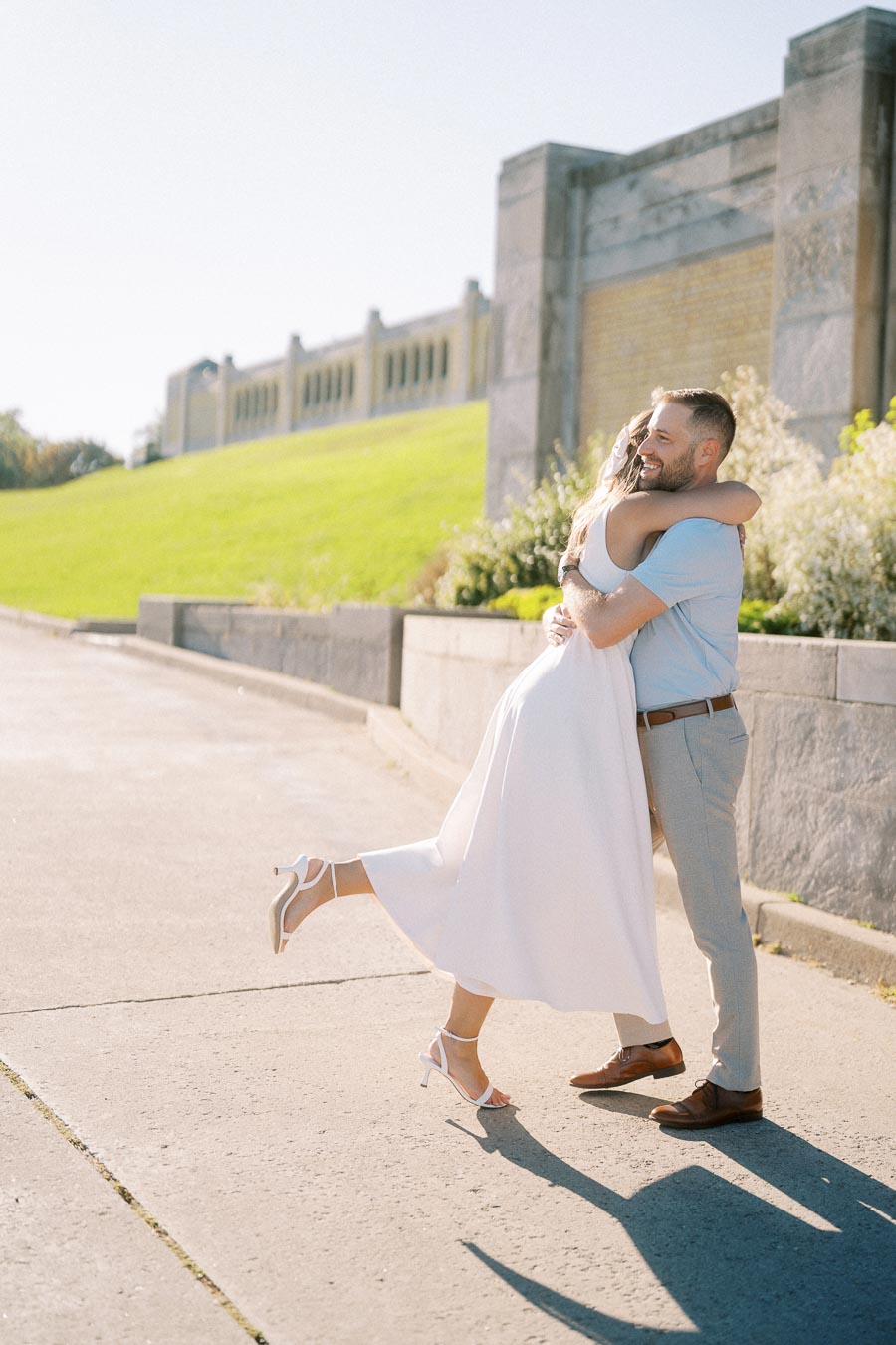 A joyful couple embracing outdoors, with the woman wearing a white dress and high heels, lifting her leg in excitement, set against a sunny backdrop featuring a stone wall and green grass.