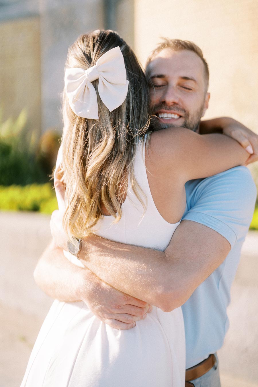 A couple embracing in a warm hug outdoors, with the woman wearing a large bow in her hair and a white dress, while the man wears a light blue shirt, both smiling in the sunlight.