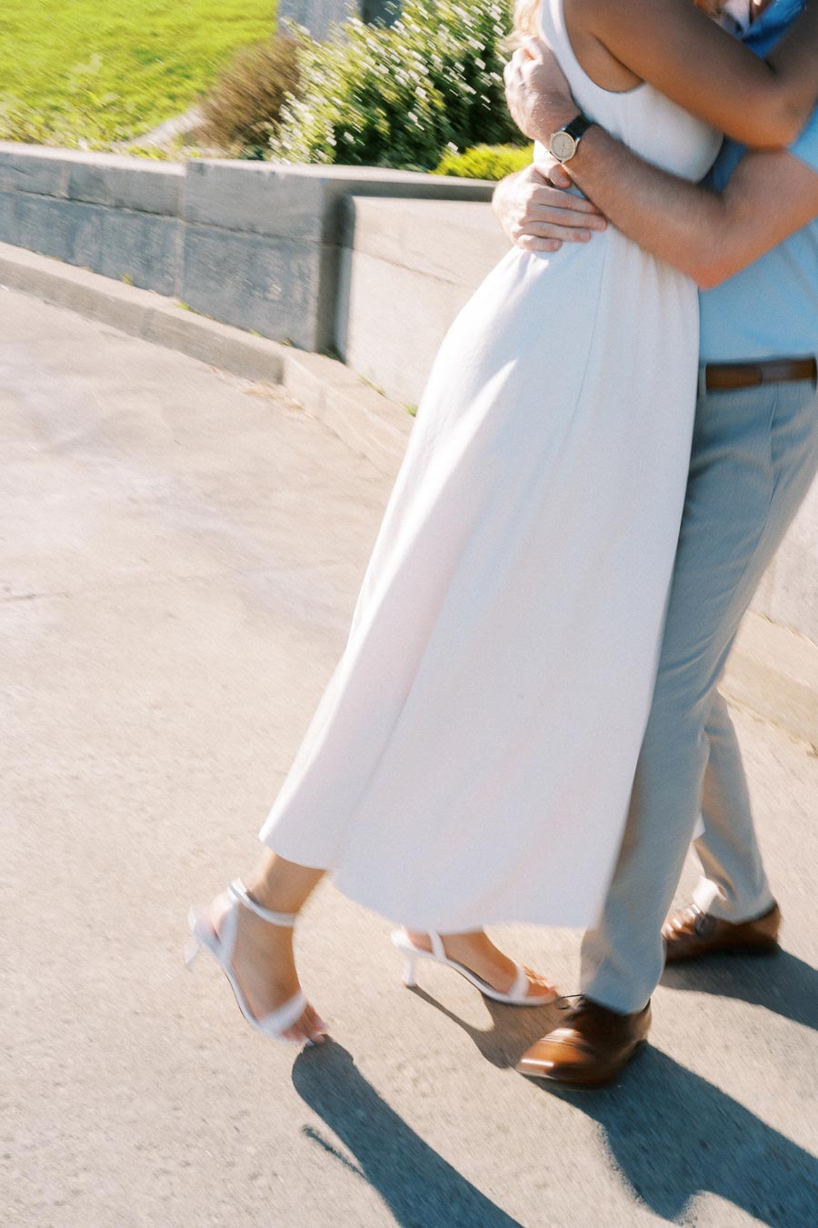 A couple embracing in a sunny outdoor setting, with the woman in a white dress and heels, and the man in light-colored pants and shoes, showcasing a romantic moment.