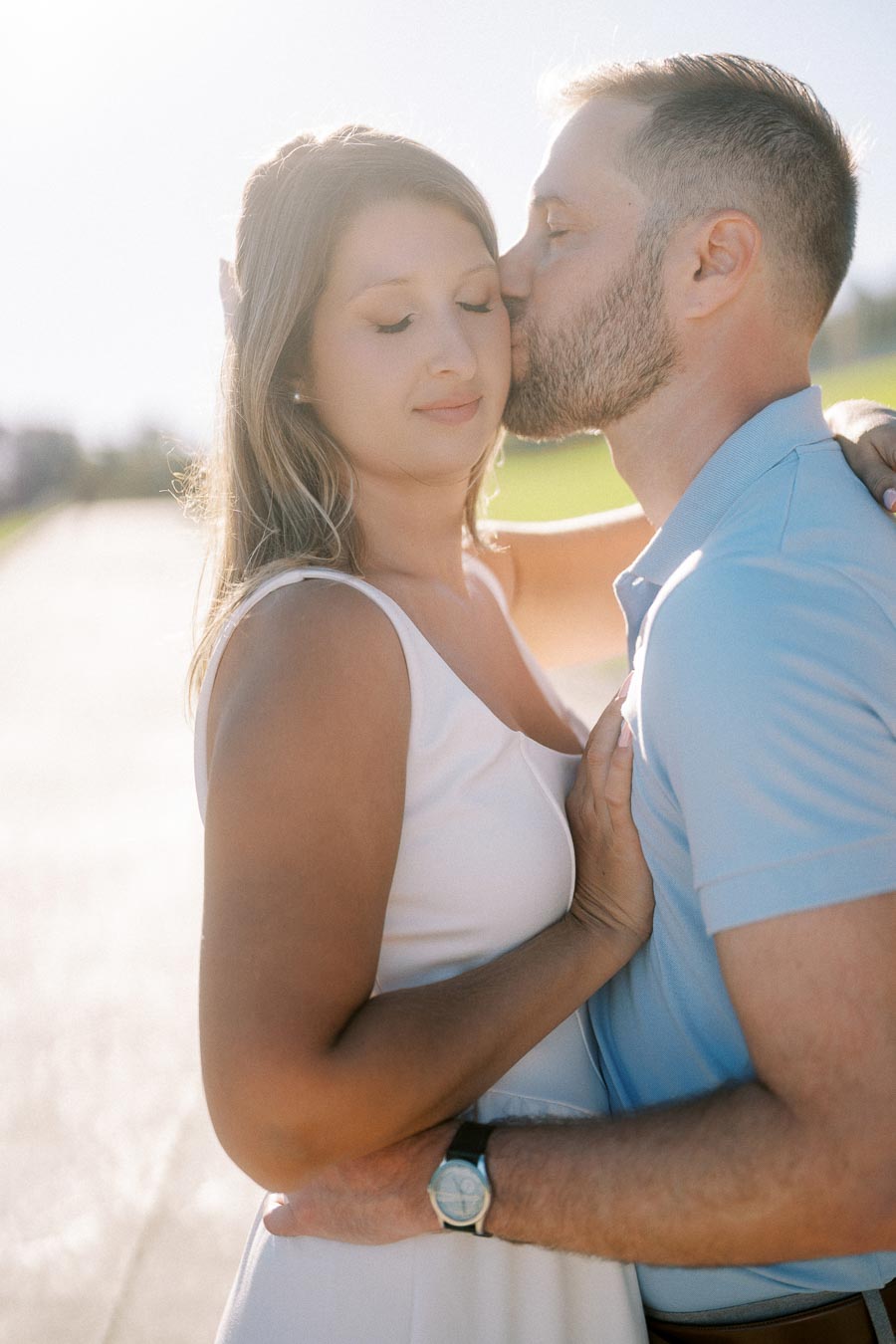 A couple embracing outdoors on a sunny day, with the man gently kissing the woman's forehead.