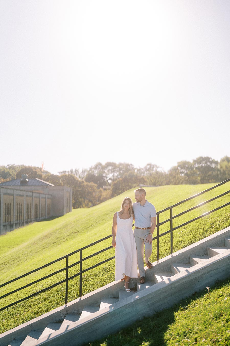 A couple walking down concrete steps on a bright sunny day, surrounded by green grass and trees in a park setting. The woman is in a white dress and the man in a light blue shirt and beige pants, both looking content and enjoying the outdoors.