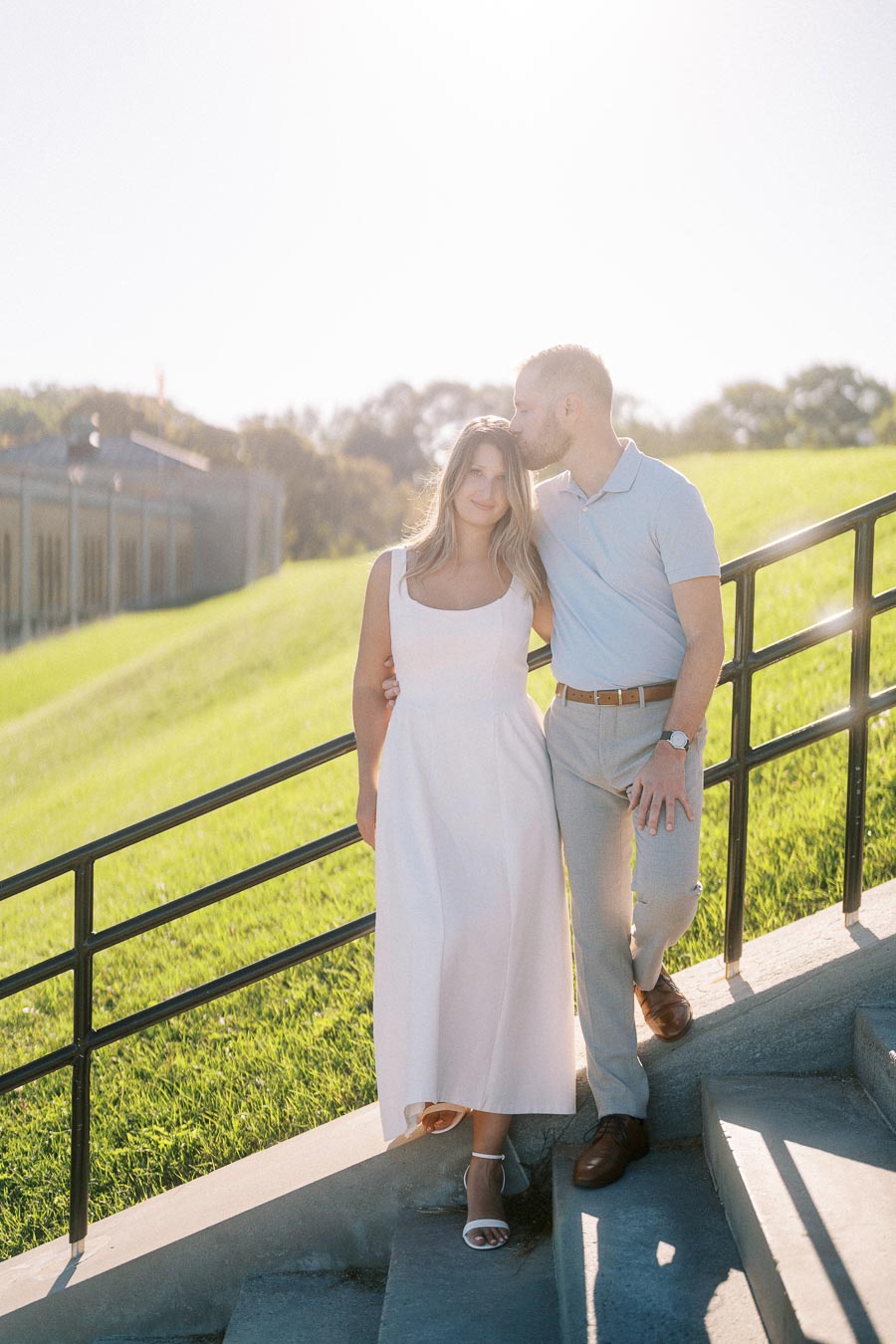 A couple enjoying a sunny day, with the man gently kissing the woman’s forehead as they stand on a staircase in a lush park setting.