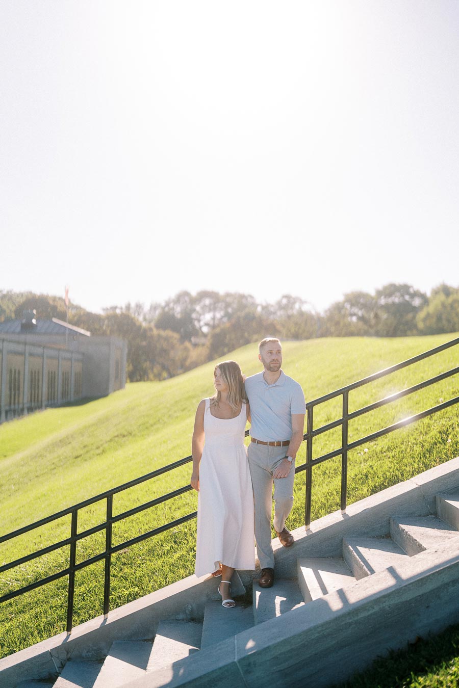 A couple walking hand in hand down a sunny outdoor staircase, surrounded by lush greenery and a building in the background.