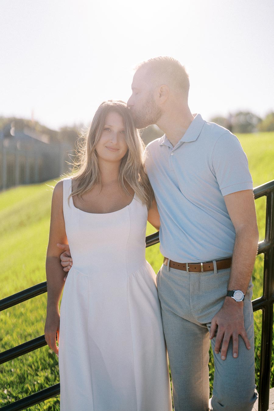 A couple standing together outdoors, with a man gently kissing a woman on the forehead. They are leaning against a fence in a sunlit field, capturing a tender and intimate moment.