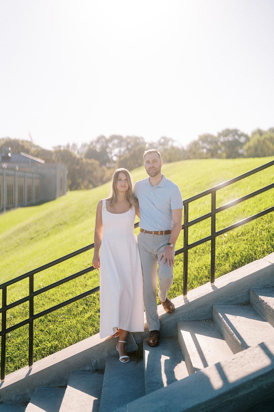 Couple standing on outdoor steps with sunlit green hill and building in background
