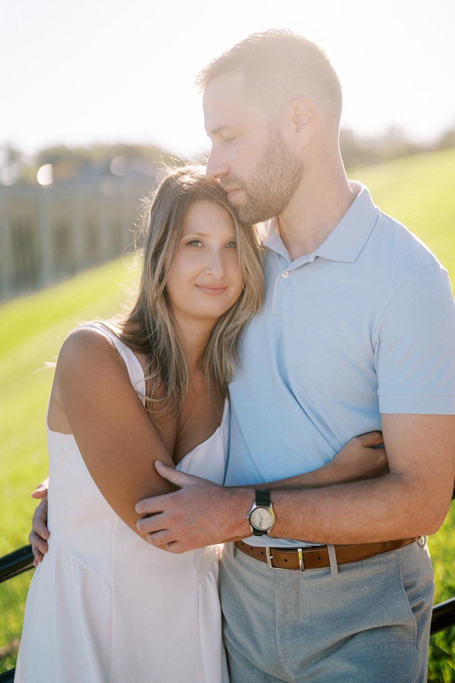 Couple embracing outdoors on a sunny day, with a woman smiling and a man gently holding her in a park setting, conveying love and serenity.