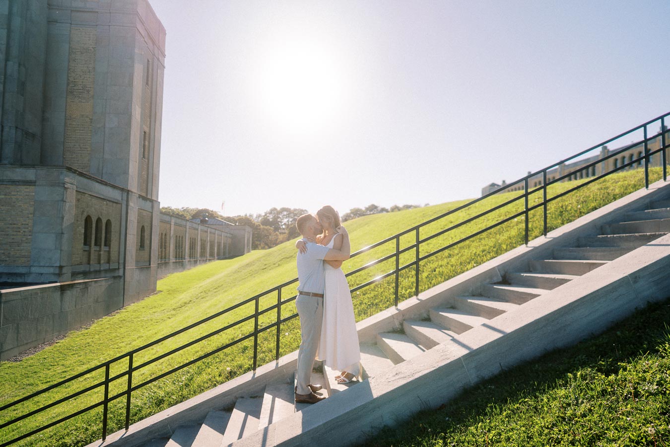 A couple embracing on a sunlit staircase beside a historic building, with green grass and clear blue sky in the background.