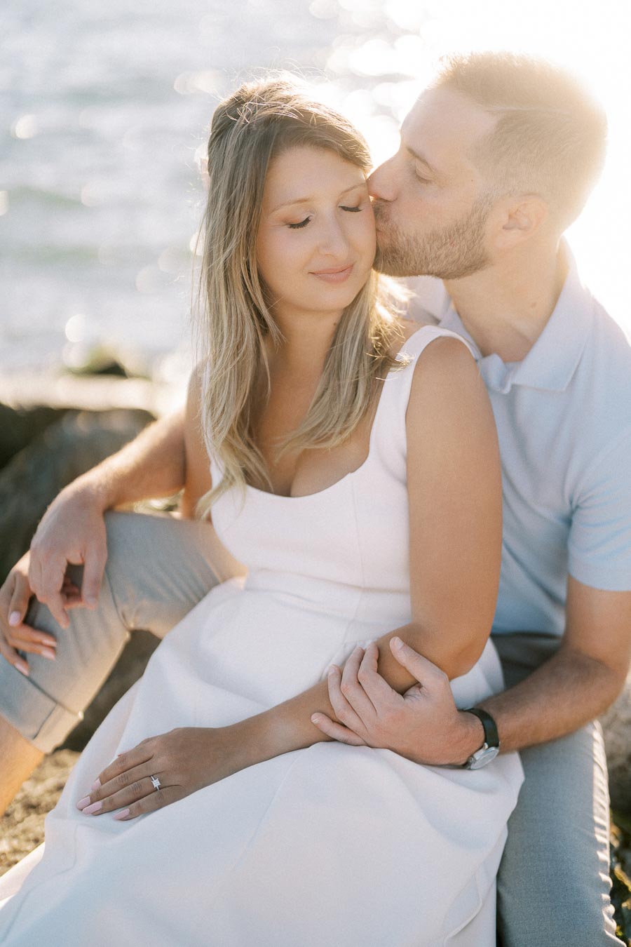 Romantic couple embracing by the water, with a man gently kissing a woman's cheek as they sit together in natural light.