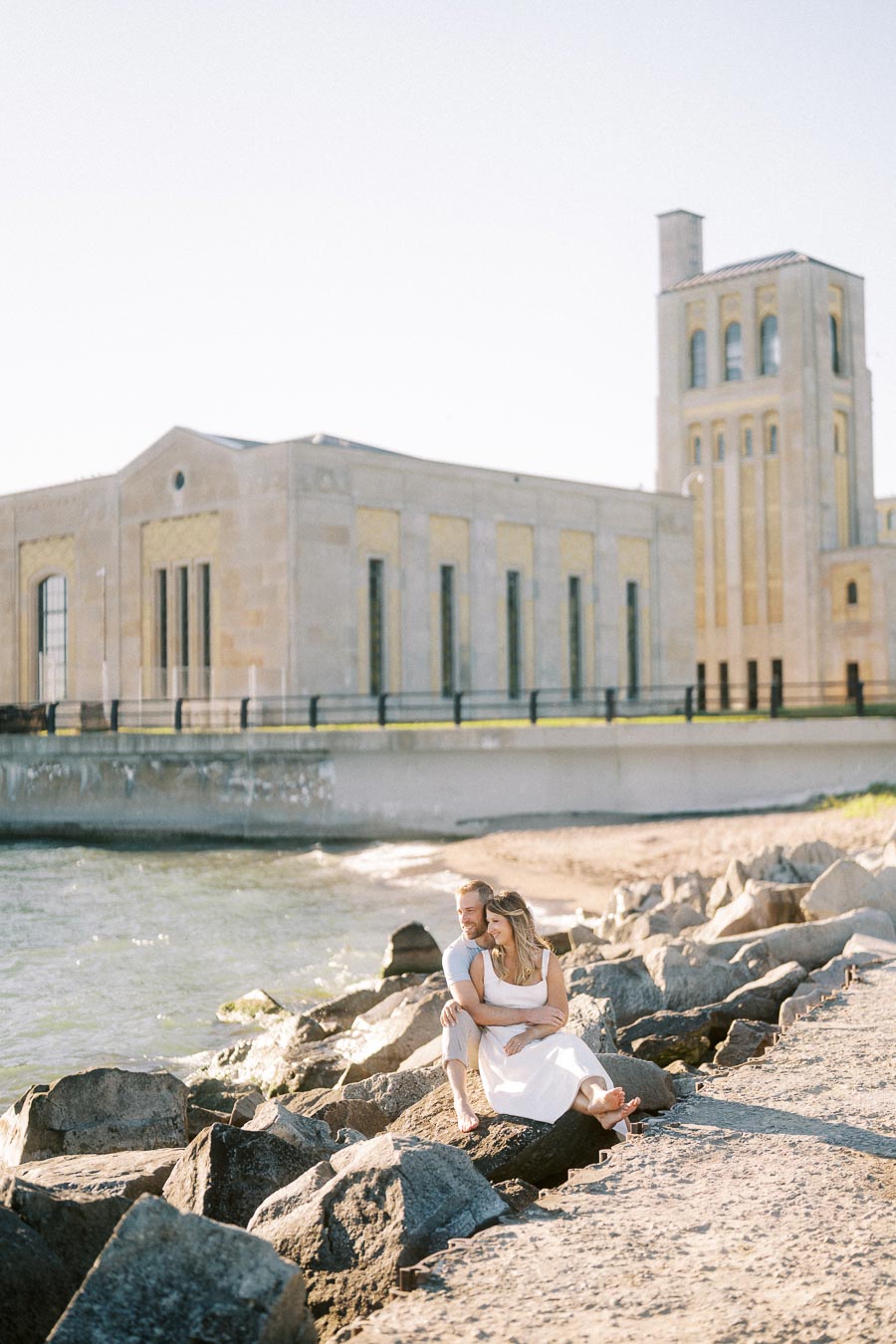 A couple sitting on rocks by the waterfront in front of an industrial-style building, embracing each other on a sunny day.
