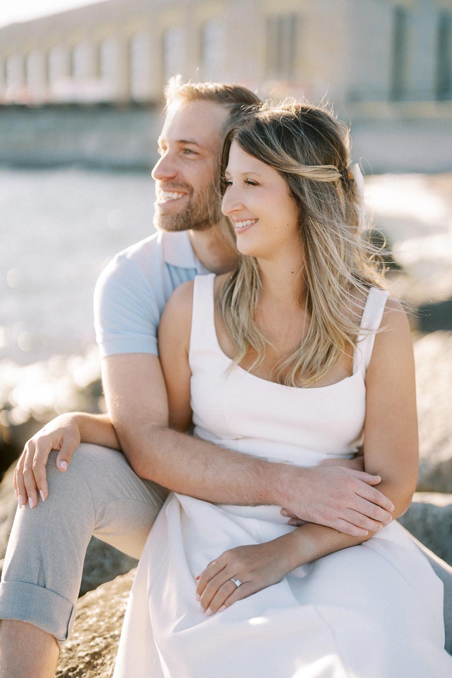 A smiling couple sitting on rocks by the waterfront on a sunny day, with a historic stone building and water in the background.