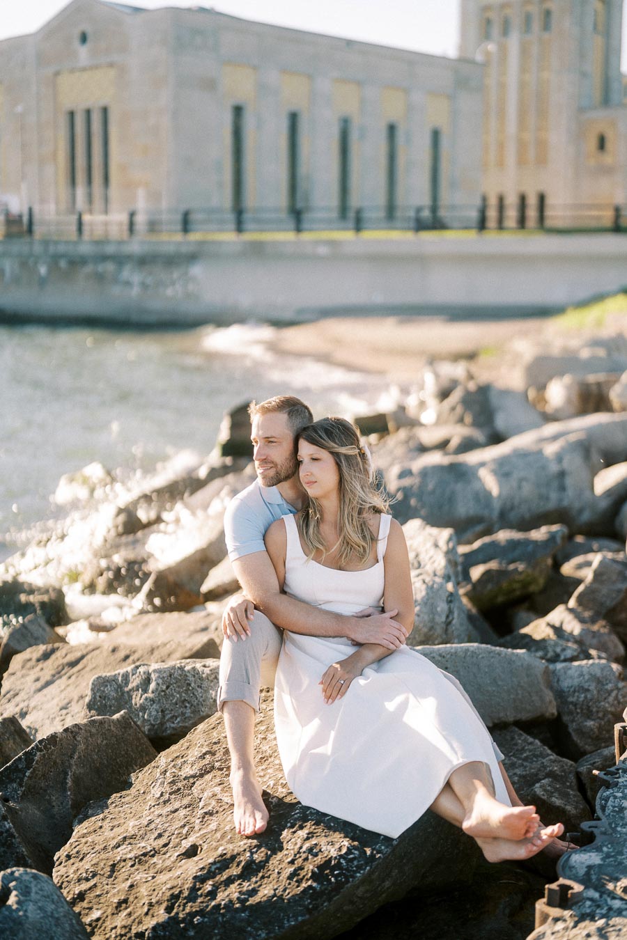 A couple sitting on rocks by the water, embracing and enjoying a peaceful moment in the sunlight with a historic building in the background.