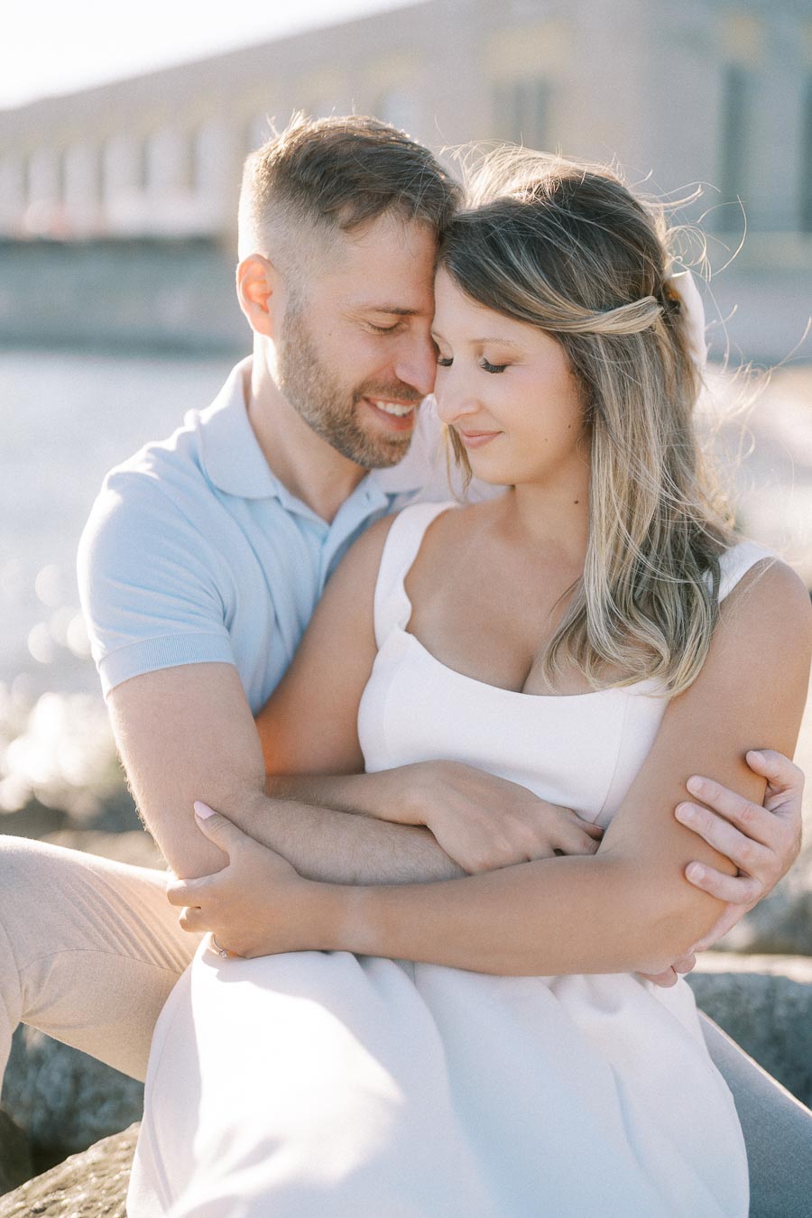 Engaged couple embracing by the waterfront, basking in sunlight during a romantic outdoor photoshoot