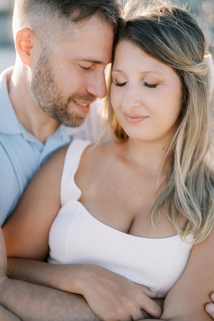 A couple embraces lovingly in soft natural light, the woman with blonde hair wearing a white dress, and the man with short hair softly leaning his forehead against hers, creating a serene, intimate moment