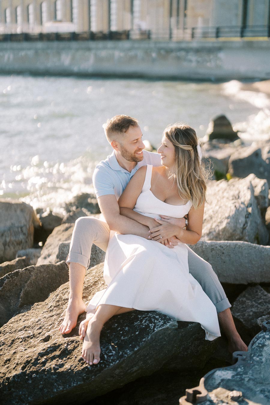 A happy couple sitting on rocks by the waterfront, embracing and smiling at each other, with a bridge and gentle waves in the background.