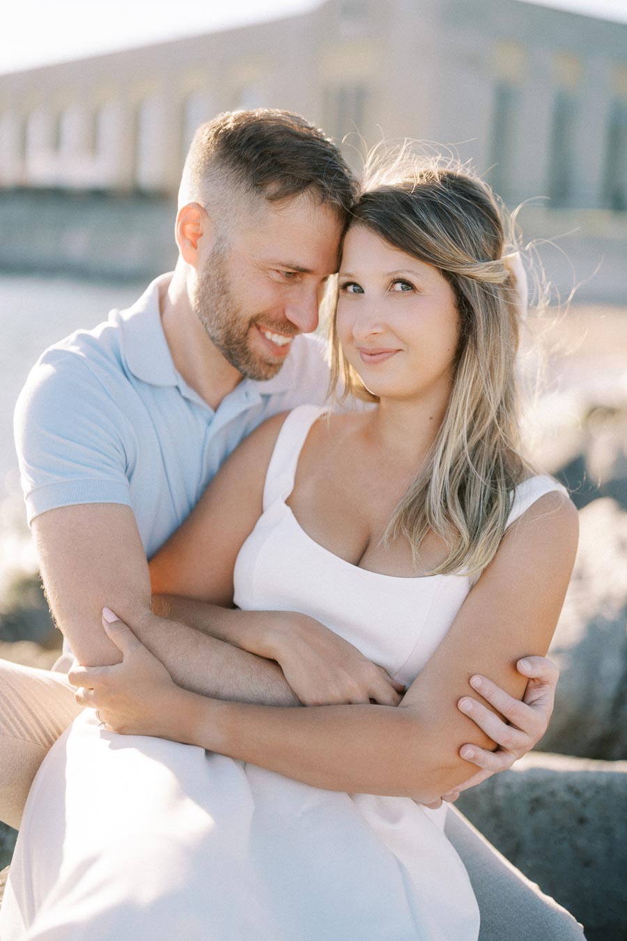 Couple embracing on a sunny day by the waterfront, man in blue shirt and woman in white dress, with a blurred architectural backdrop