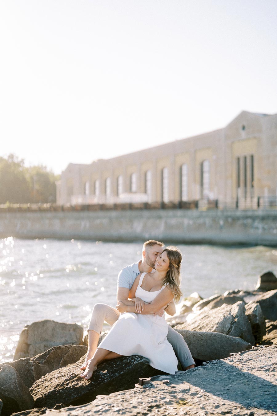 A couple sitting on rocks by the water, embracing and smiling under a clear sky. The background features a historical stone building with tall windows.