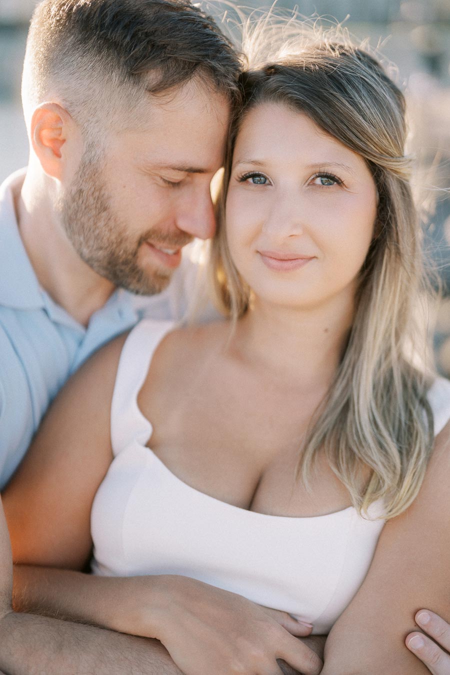 Couple embracing in a warm, intimate moment, with the woman looking at the camera and the man gently leaning in, both bathed in soft natural light.
