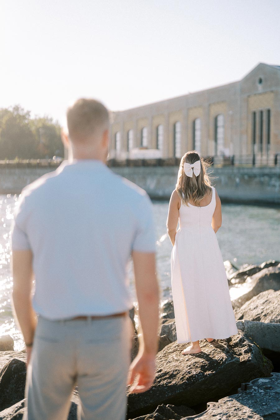Man in light blue shirt standing on rocks by a waterfront, facing a woman in a white dress with a large bow in her hair, as an industrial-style building and trees are visible in the background.