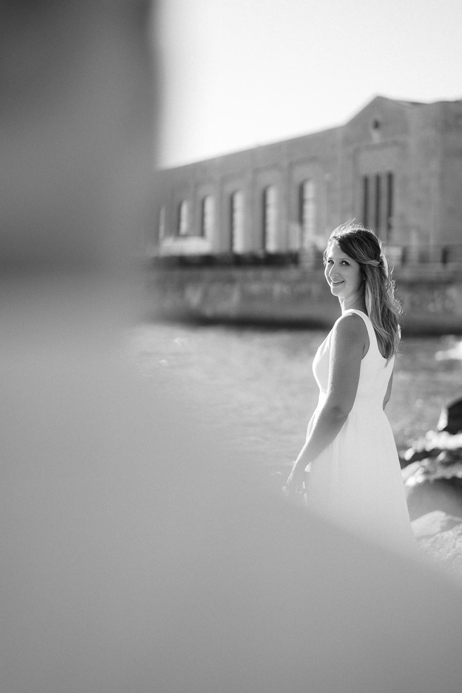 Black and white image of a woman in a white dress smiling by the waterfront with an industrial building in the background.
