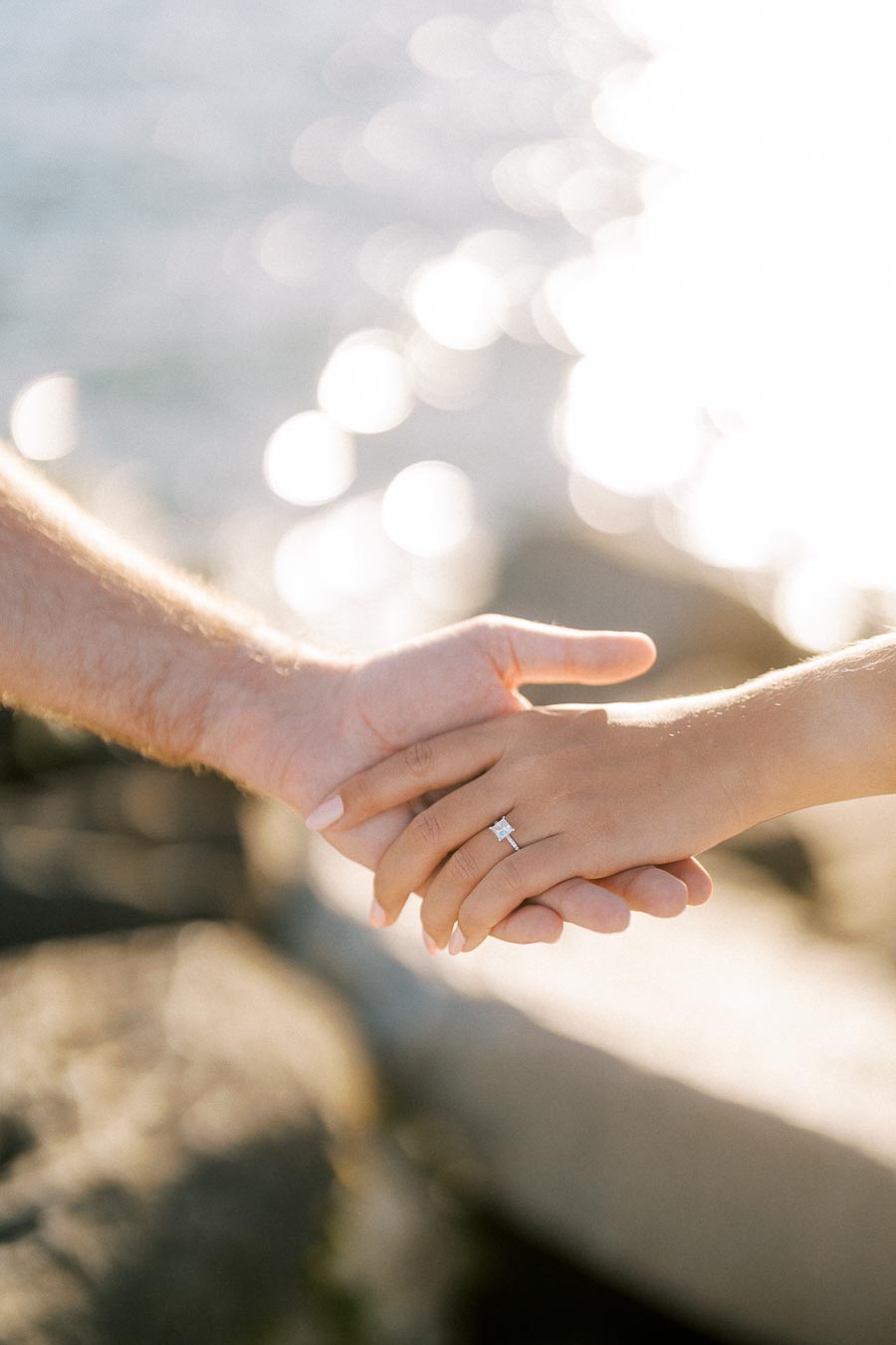 Couple holding hands with a diamond engagement ring by the ocean, sunlight sparkling on the water