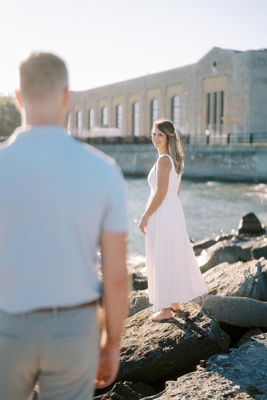 A woman in a white dress stands on rocks by the water, looking back at a person in a light blue shirt. The setting is sunny with an industrial building in the background.