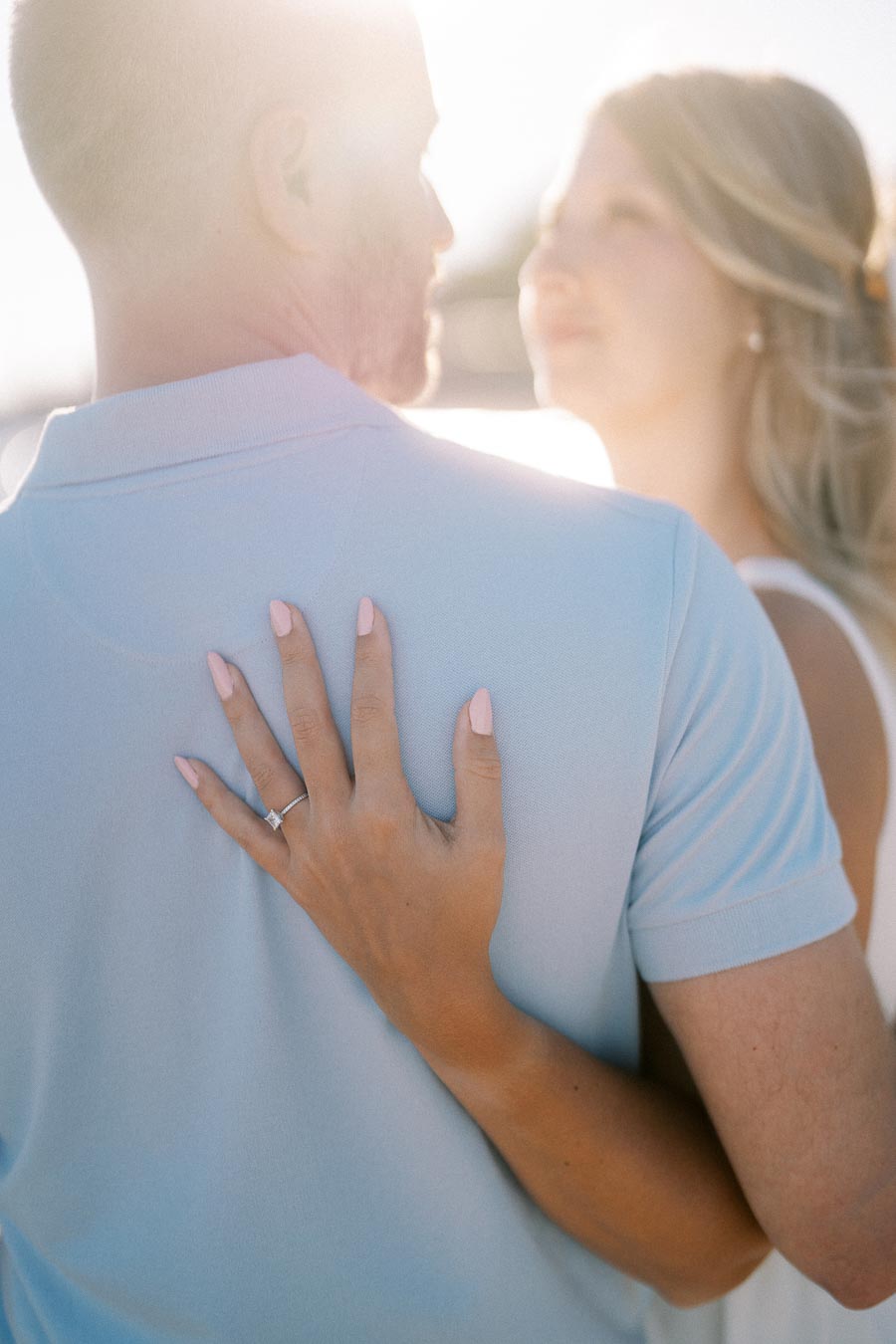 A close-up of a woman's hand with a diamond engagement ring resting on a man's shoulder, both bathed in soft sunlight, symbolizing love and commitment.